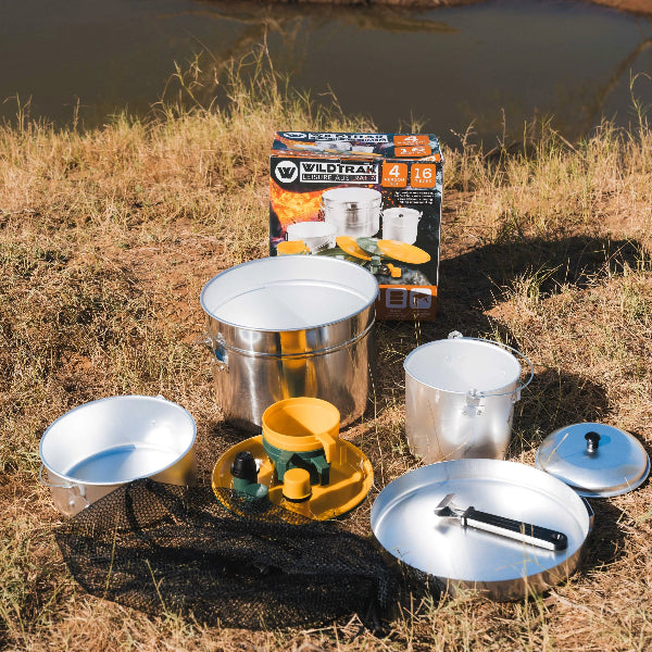 Camping cookware set with pots and pans on a grassy area near a body of water.