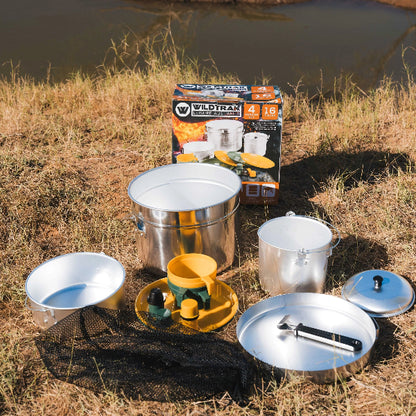 Camping cookware set with pots and pans on a grassy area near a body of water.